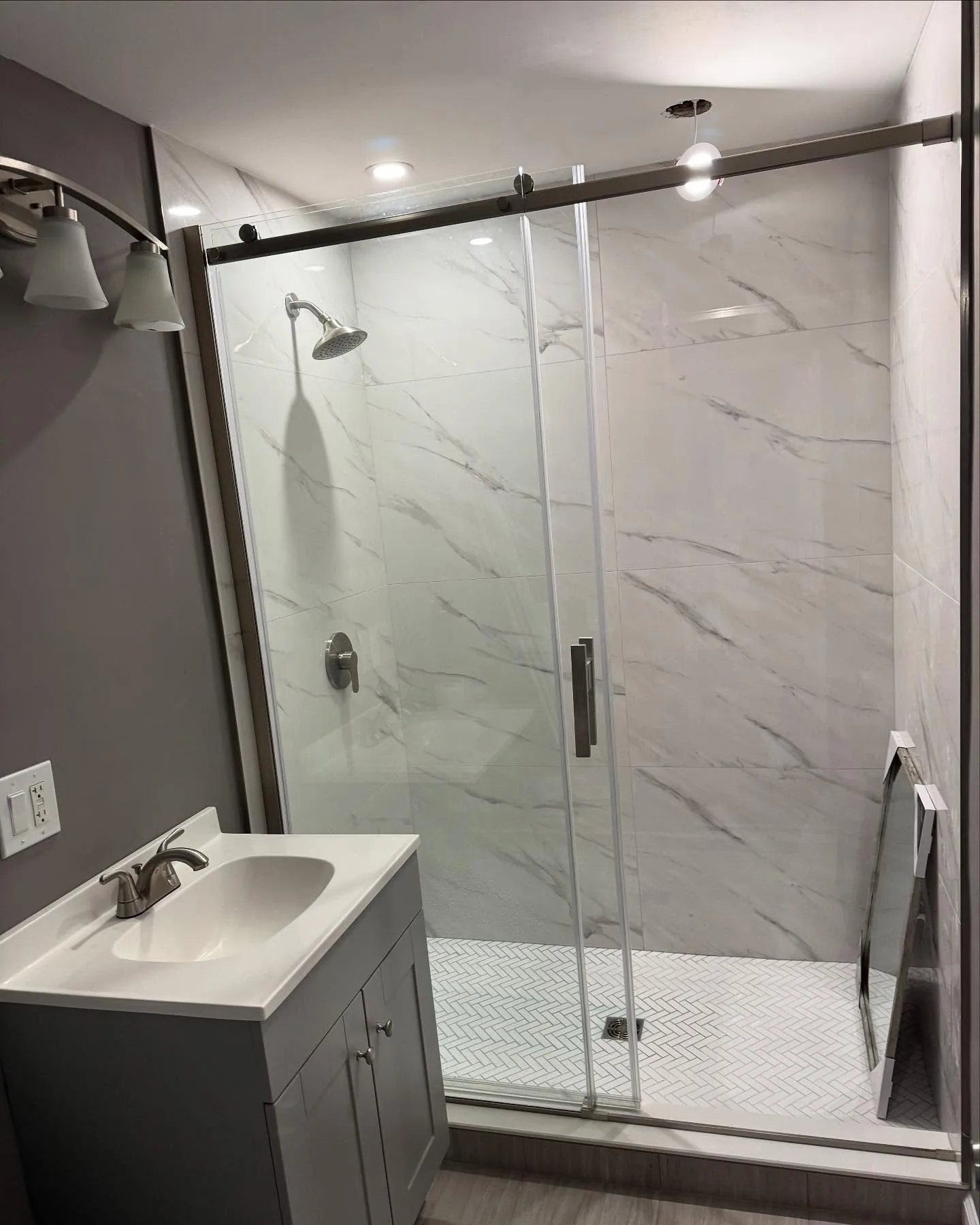 View of a completed custom tile bathroom remodel featuring a glass-enclosed shower with marble-patterned walls and a grey vanity cabinet.